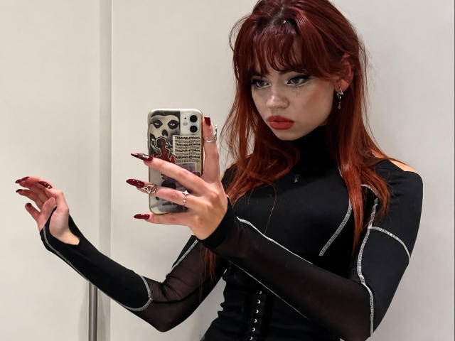 YettaGlumac mit roten Haaren, einem schwarzen Langarmshirt und roten Nägeln, hält ein Telefon, zeigt ihre langen Fingernägel, blickt in einen Spiegel, in einem Badezimmer..