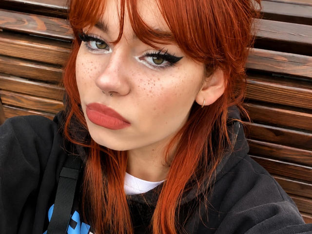 YettaGlumac hat rotes Haar, Sommersprossen, grüne Augen und trägt auffälligen Eyeliner mit langen Wimpern. Sie trägt einen Nasenring und dunklen Lippenstift. Sie ist in einer schwarzen Kapuzenjacke gekleidet..
