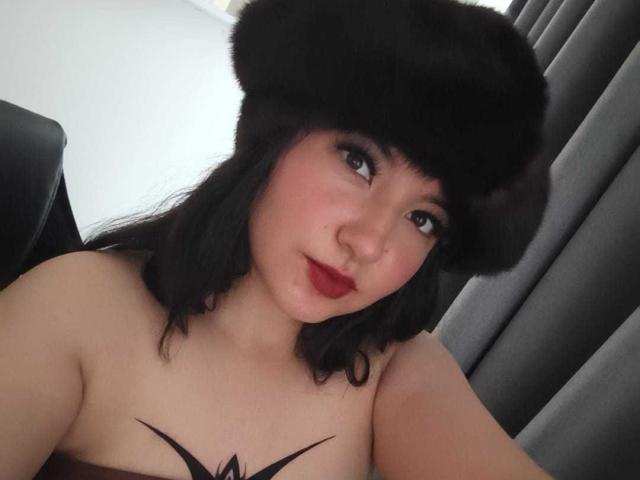 Susan Monroy, com cabelo escuro, batom vermelho e um chapéu de pele preta, exibindo os ombros nus e uma tatuagem preta abaixo dos seios. Ela está sentada, olhando para a câmera e sorrindo levemente..