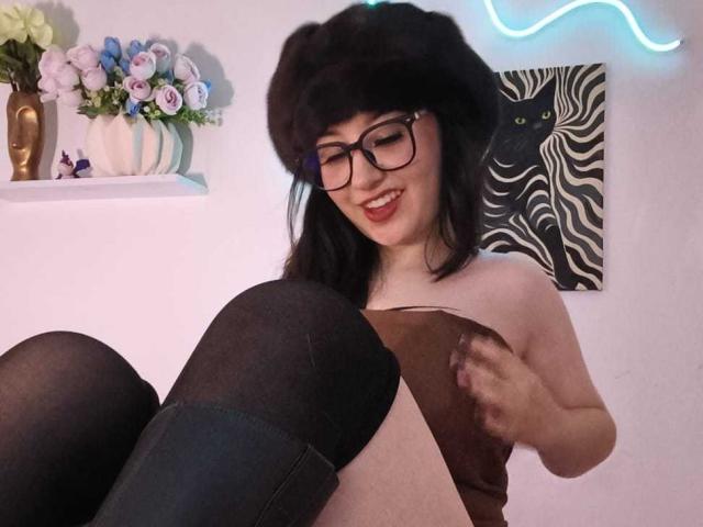 SusanMonroy met een zwarte bontmuts, een bril en een glimlach, die een bruin boek vasthoudt, draagt zwarte kniekousen en een bruin topje, waarbij haar borsten zichtbaar zijn, in een gezellige kamer met blauw neonlicht..