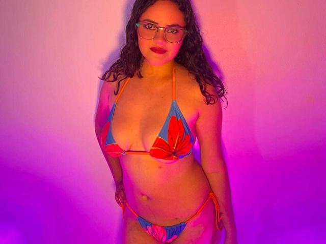 EmilyColsson in a colorful bikini, showing her tits in a red and blue floral top, and matching red and blue floral panties, with curly hair and glasses.