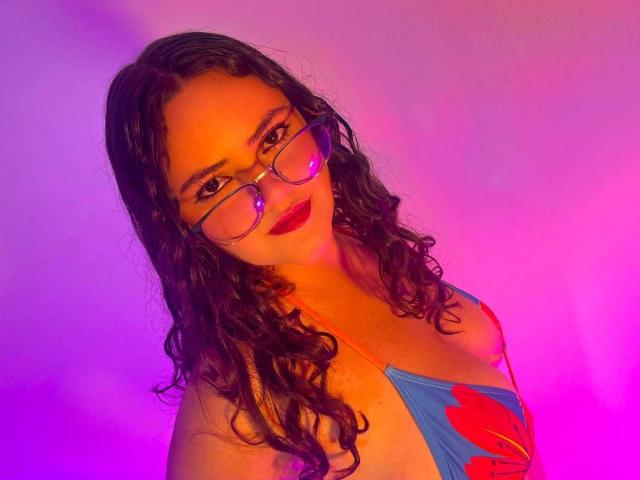 Emily Colsson, con cabello negro rizado, gafas y lápiz labial rojo, vestida con un top de bikini azul y rojo, sonriendo a la cámara bajo una iluminación morada y rosa..