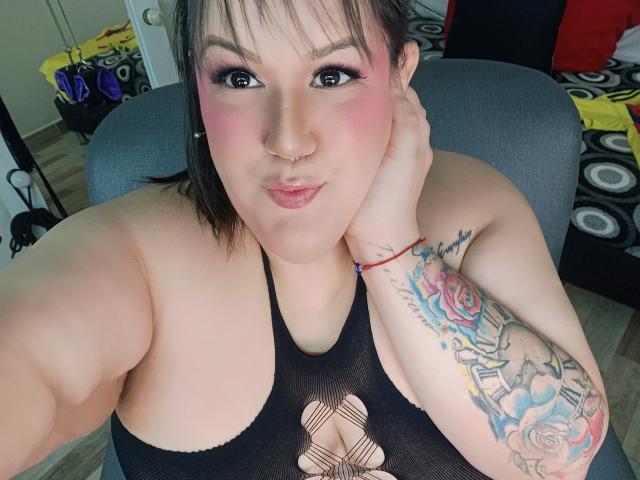 GiiannaMorrys, avec un blush rose, un haut en maille noire, un anneau au nez, un bras tatoué et un rouge à lèvres rose, tenant son menton, montrant ses seins et une expression mignonne et sexy..