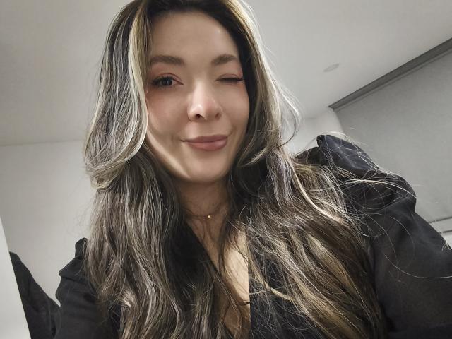 TaniaSandersX mit zwinkerndem Auge, langem, grau- und blond gefärbtem Haar, lächelnd, trägt ein schwarzes Hemd, eine goldene Halskette, hat helle Haut und dezentes Make-up, auf einem Selfie-ähnlichen Foto..
