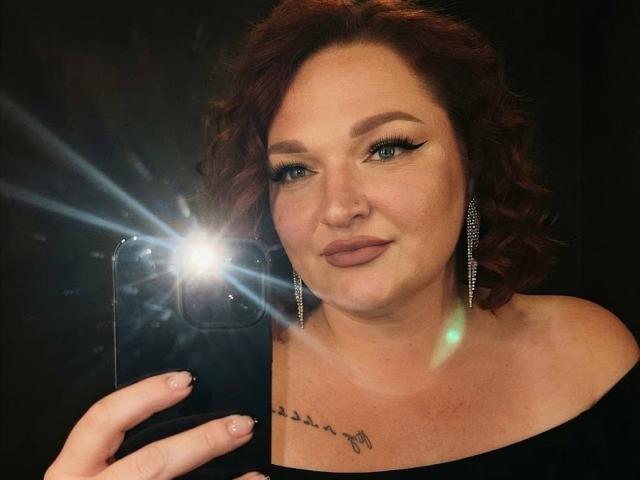 AngelQSky with red curly hair, wearing black off-shoulder top, holding phone with bright flash, showing tattoo under neck, wearing silver earrings, smiling subtly, blue eyeliner.