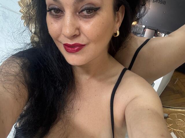 KattSuunny has dark hair, red lipstick, black eyeliner, gold hoop earrings, and a black spaghetti-strap top, showing her tits and a confident smile in a selfie.