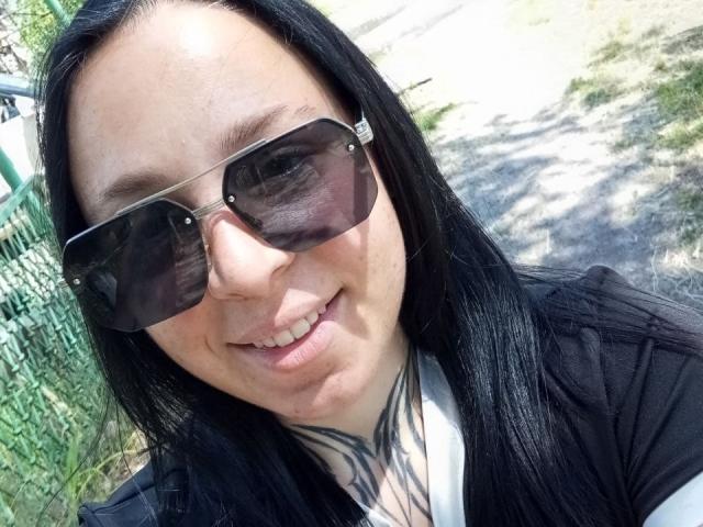 MillaSouReety, com cabelo preto, usando óculos de sol, sorrindo, tem tatuagens no pescoço e está vestida com uma blusa preta e branca. Seu nariz é ligeiramente arrebitado..