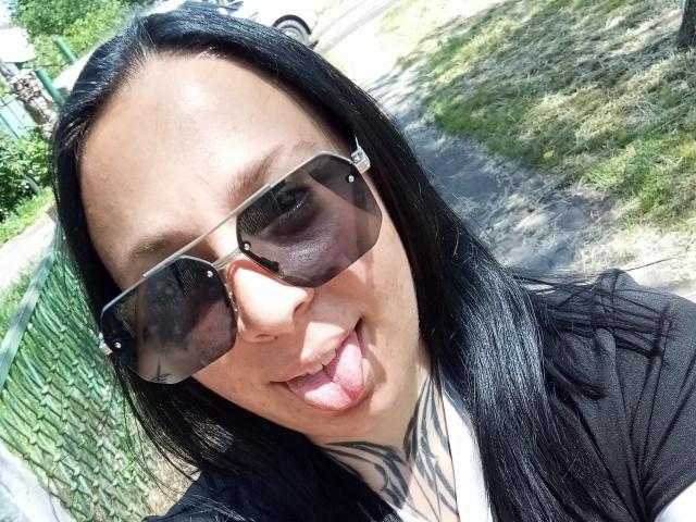 MillaSouReety, com cabelo preto, óculos de sol marrons e uma tatuagem no pescoço, está de língua para fora. Ela está vestindo uma blusa preta. Suas nádegas não estão visíveis..