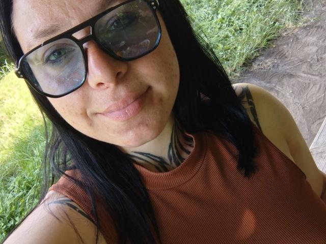MillaSouReety, com óculos pretos, regata marrom, tatuagens pretas no pescoço e um sorriso discreto, mostrando sardas e cabelos longos e pretos em um selfie..