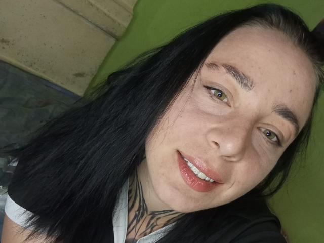 MillaSouReety tiene el pelo largo y negro, ojos verde claro y un tatuaje en el cuello. Está sonriendo con los labios rojos, mostrando los dientes blancos, y tiene una pequeña peca en la nariz..