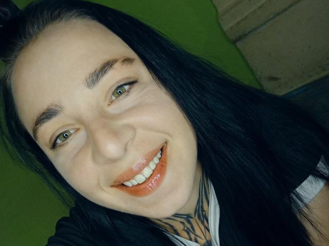 MillaSouReety, com pele clara, cabelo preto, olhos verdes e uma tatuagem no pescoço, sorrindo com seus lábios laranja brilhantes. Suas sobrancelhas pretas se destacam contra seus dentes brancos..