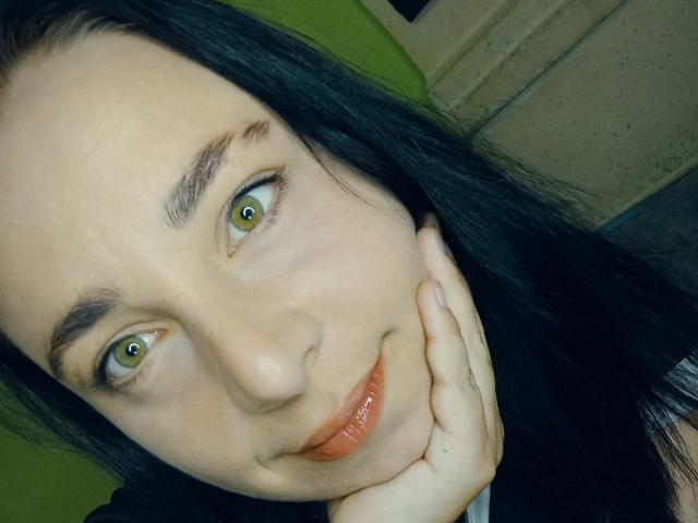 MillaSouReety, com olhos verdes, cabelo preto e lábios alaranjados brilhantes, apoiando o queixo na mão, sorrindo levemente. Suas sobrancelhas são escuras e bem definidas..