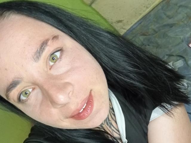 O rosto de MillaSouReety, com olhos verdes, cabelo preto e liso, pele clara e lábios vermelhos, vestindo uma camisa preta e branca, sorrindo levemente. Suas nádegas não estão visíveis..