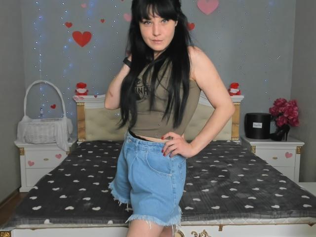 WenTina with long black hair, green sleeveless top, and frayed blue denim shorts, standing in bedroom, hand on hip, looking at camera, with a confident expression.