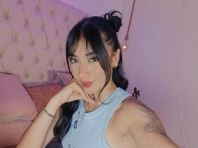 KatheLeclecr mit dunklem Haar zu einem Dutt gebunden, trägt ein blaues Tanktop, lächelt, berührt ihre roten Lippen, hat ein Tattoo am rechten Arm, im Hintergrund ein pinkes Kissen..