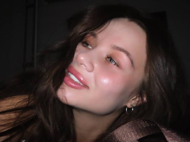 Azzie Stelman, con la pelle luminosa, gli occhi verdi e le labbra rosa, sorride leggermente con i capelli castano scuro che le sventolano di lato, indossando un orecchino a cerchio e una camicia a quadri marrone..