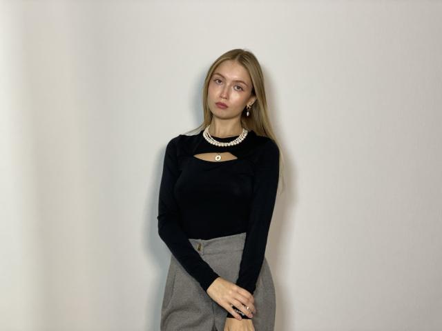 SherlyTedford mit blonden Haaren, einem schwarzen Oberteil mit langen Ärmeln, einem grauen Rock mit hoher Taille, einer Perlenkette und Ohrringen, steht vor einer schlichten Wand, blickt leicht zur Seite und hält die Hände zusammen..
