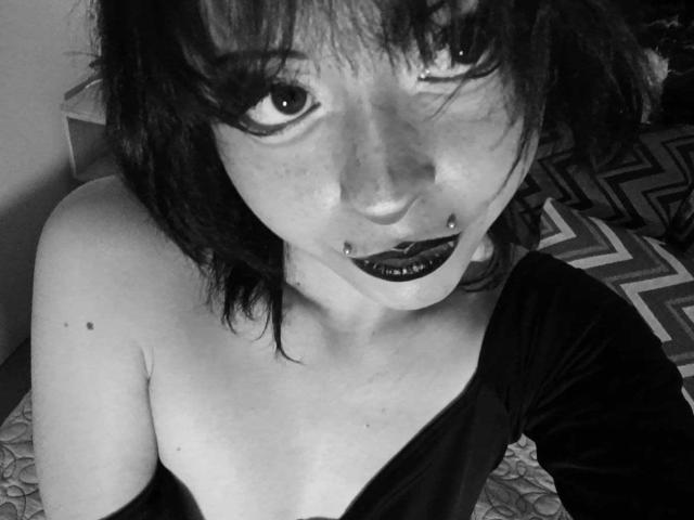 ViolettClay with dark hair, black lipstick, and a lip piercing, wearing an off-shoulder top, looking up at the camera, showing her bare shoulders and a small mole on her chest.