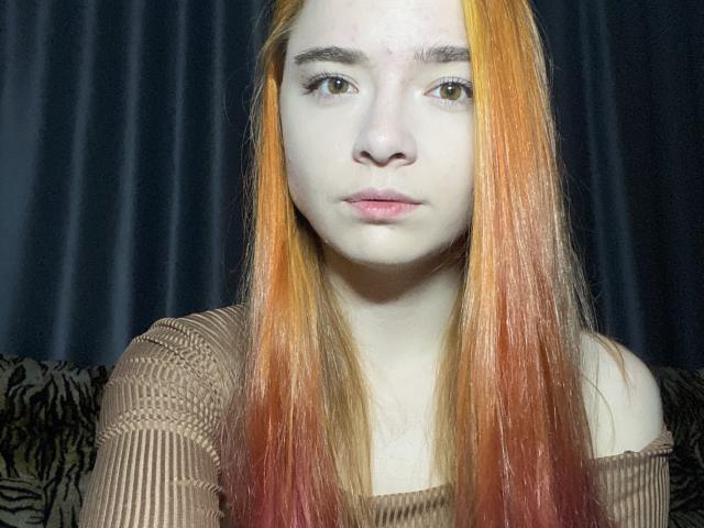 MarlanaBub ha la pelle chiara, lunghi capelli con sfumatura dal rosso all'arancione, occhi marroni e indossa una maglia marrone a coste, con una spalla scoperta. Ha un'espressione seria ed è al centro dell'immagine..