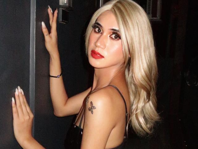 Precious, met blond haar, rode lippenstift en een zwarte tatoeage op haar schouder, staat tegen een donkere muur en toont haar achterwerk, met haar rug naar de camera gericht..