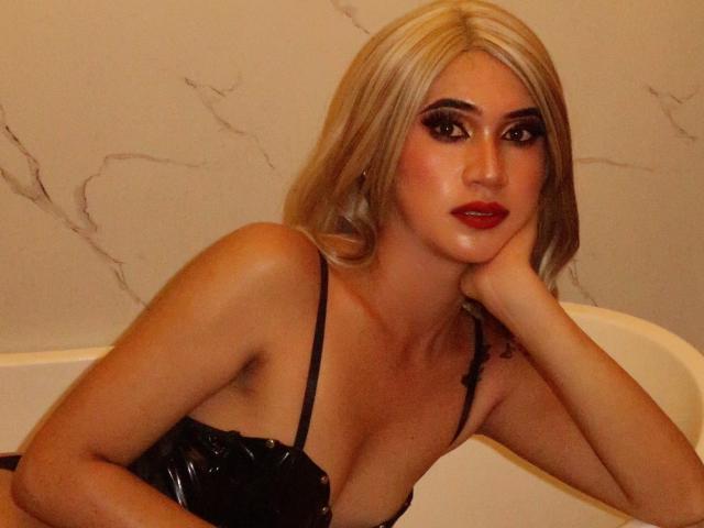 Precious, met blond haar, rode lippenstift en zwarte lingerie, leunt met haar hoofd op haar hand en laat haar borsten zien in een wit bad. Haar make-up is opvallend en aantrekkelijk..