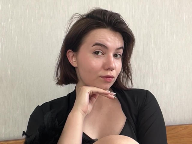 Elina Haycock mit kurzen, braunen Haaren, voller Lippen und einem Nasenring, trägt ein schwarzes Oberteil, legt ihr Kinn in die Hand und zeigt ihre Brüste, in einer entspannten Pose..