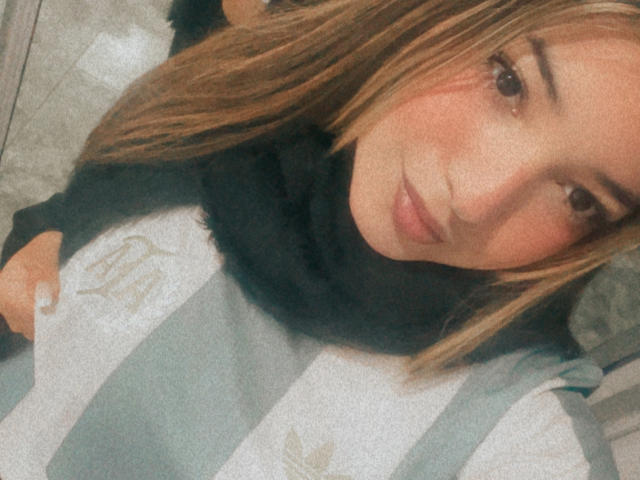 AmbarDunn, con i capelli castano chiaro, una sciarpa di pelliccia nera e un maglione grigio a righe, sorride dolcemente alla fotocamera, mostrando il suo viso carino e il trucco leggero..