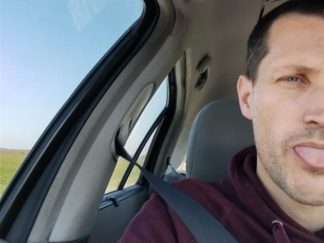 Il volto di MisterFredX, con capelli castano corti, occhi azzurri e una leggera barba, con la lingua fuori, indossa una felpa color borgogna, con la cintura di sicurezza allacciata, all'interno di un'auto con un finestrino sul lato sinistro..