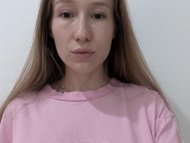 Kareen Brumbaugh, mit langen braunen Haaren, trägt ein pinkes Hemd, hat einen neutralen Gesichtsausdruck und eine kleine Pickel auf ihrer linken Wange, vor einer weißen Wand..