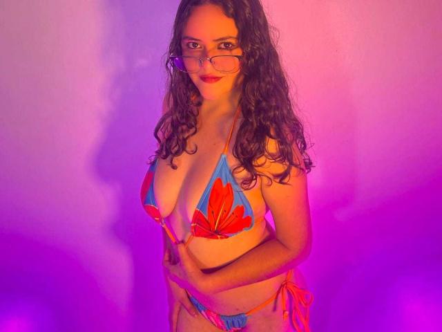 EmilyColsson with curly brown hair, glasses, and red lipstick, wearing a blue bikini with red seashell patterns, holding her tits, standing in pink and purple lighting.