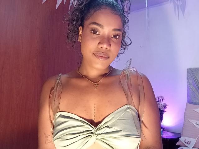 Caylin, con cabello rizado, piel de tono marrón medio, vestida con un vestido de satén verde con tirantes transparentes, maquillaje discreto, un collar dorado y un tatuaje en su brazo izquierdo, mirando a la cámara..