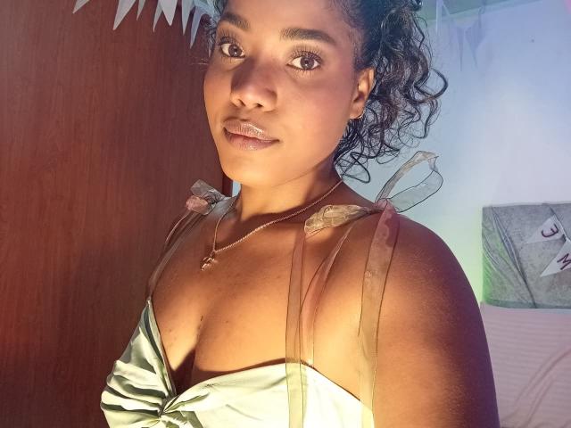 Caylin, com cabelos cacheados escuros, pele morena e seios pequenos, vestindo um vestido verde brilhante com alças transparentes, sorrindo para a câmera, usando um colar de ouro..