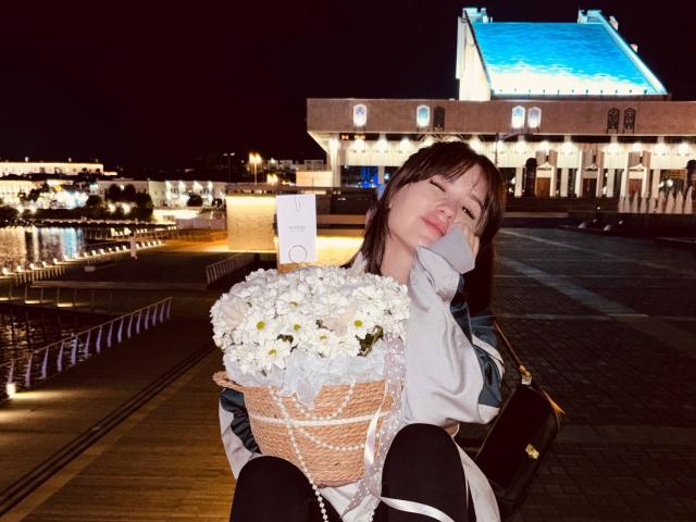 Anna Bauknight con flores blancas en una cesta de mimbre, vistiendo una camisa blanca, pantalones negros y una chaqueta verde, sentada por la noche, sosteniéndose la cara con una expresión de cansancio..