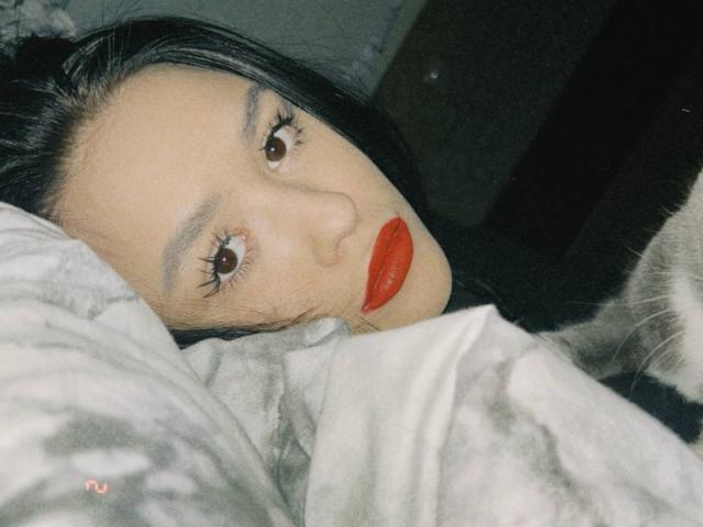 Anna Bauknight, con rossetto rosso, capelli neri e occhi marroni, sdraiata su un letto con lenzuola grigie, il viso leggermente rivolto di lato, che guarda la fotocamera..