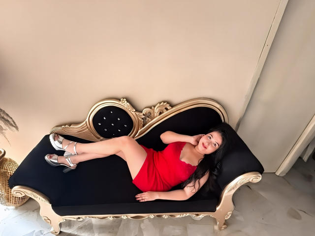 BbwLuxuryMuse ligt op een versierde zwarte en gouden bank, gekleed in een rode jurk en zilveren hoge hakken, waarbij haar billen en borsten zichtbaar zijn, en ze heeft een schattige glimlach..