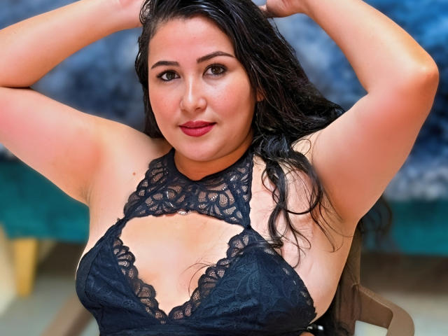 BbwLuxuryMuse met nat zwart haar, armen omhoog, gekleed in een zwart kant topje dat haar grote borsten onthult, rode lippenstift en een lichte glimlach, zittend in een stoel..