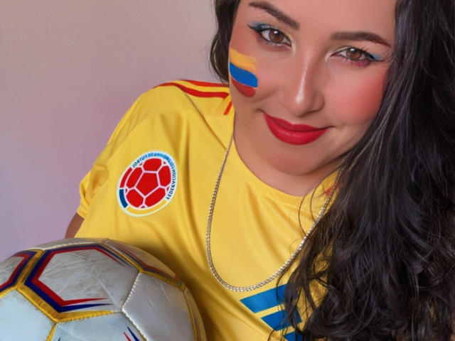 BbwLuxuryMuse avec rouge à lèvres rouge, maquillage facial bleu et jaune, t-shirt de foot jaune, tenant un ballon de foot, cheveux noirs longs et collier en argent, souriant à la caméra..