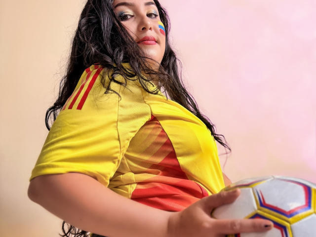 BbwLuxuryMuse avec de longs cheveux noirs, un maillot de foot jaune, des rayures rouges et jaunes, tenant un ballon de foot, du maquillage bleu sur la joue, regardant vers le bas, fond rose. Seins visibles sous le maillot..