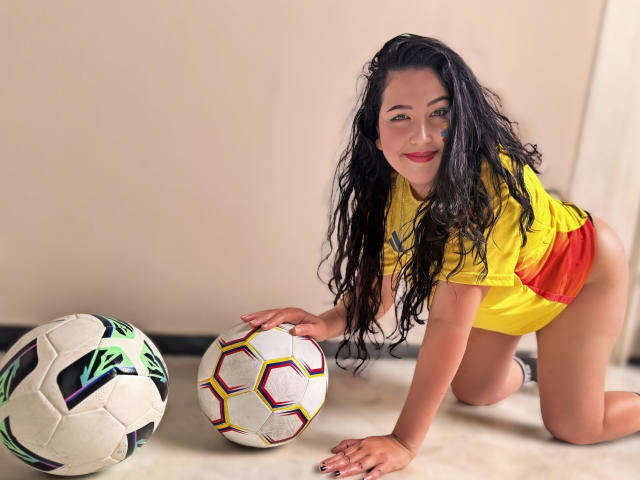BbwLuxuryMuse, with long wet black hair, kneels on floor, wearing yellow and red soccer jersey, touching soccer balls, smiling, showing her ass, in bright indoor setting.