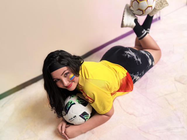 BbwLuxuryMuse lying on her stomach, wearing a yellow and red shirt, black shorts, and white soccer shoes, holding a soccer ball, with blue face paint, and curly black hair.