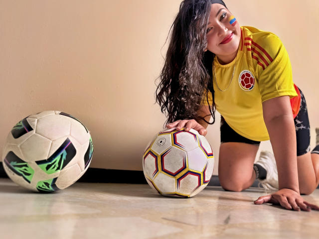 BbwLuxuryMuse à genoux, portant un maillot de foot jaune, un short noir, avec de longs cheveux bouclés, du maquillage bleu sur le visage, tenant un ballon hexagonal blanc, et un deuxième ballon de foot avec des graffitis verts sur la gauche..