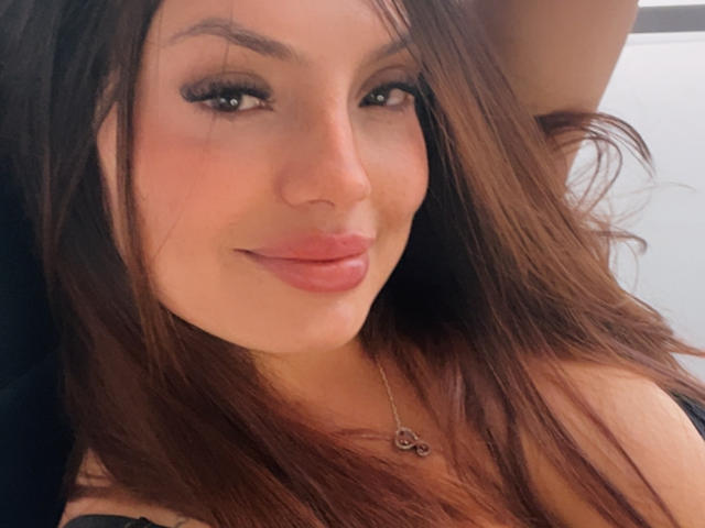 GalaMalkova ha i capelli castani, labbra piene rosate e un sorriso adorabile, indossa una maglietta nera con una delicata collana. Guarda la fotocamera con un'aria rilassata e sicura di sé..