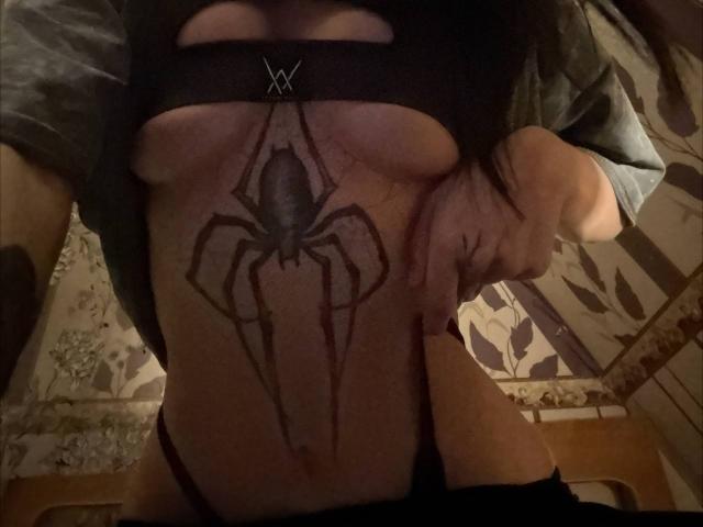 Le ventre nu de ShylaCatillo révèle un tatouage de toile d'araignée noire juste en dessous de ses seins. Elle porte un haut noir avec des découpes et a sa main posée sur sa hanche droite..