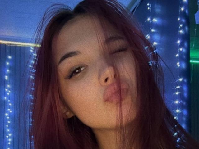 ShylaCatillo, con le labbra piene, che fa l'occhiolino e con i capelli rossi, con ciocche che le coprono il viso, che indossa un sottile trucco per gli occhi, sullo sfondo di lucine blu, appare carina e giocosa..