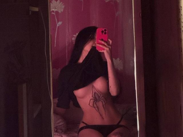 ShylaCatillo mit schwarzen Haaren, die ihr Hemd hochhebt, um ihre kleinen Brüste, schwarze Unterwäsche und ein Spinnentattoo auf ihrer Brust zu zeigen, macht ein Selfie mit einem rosa Handy vor einem Spiegel..