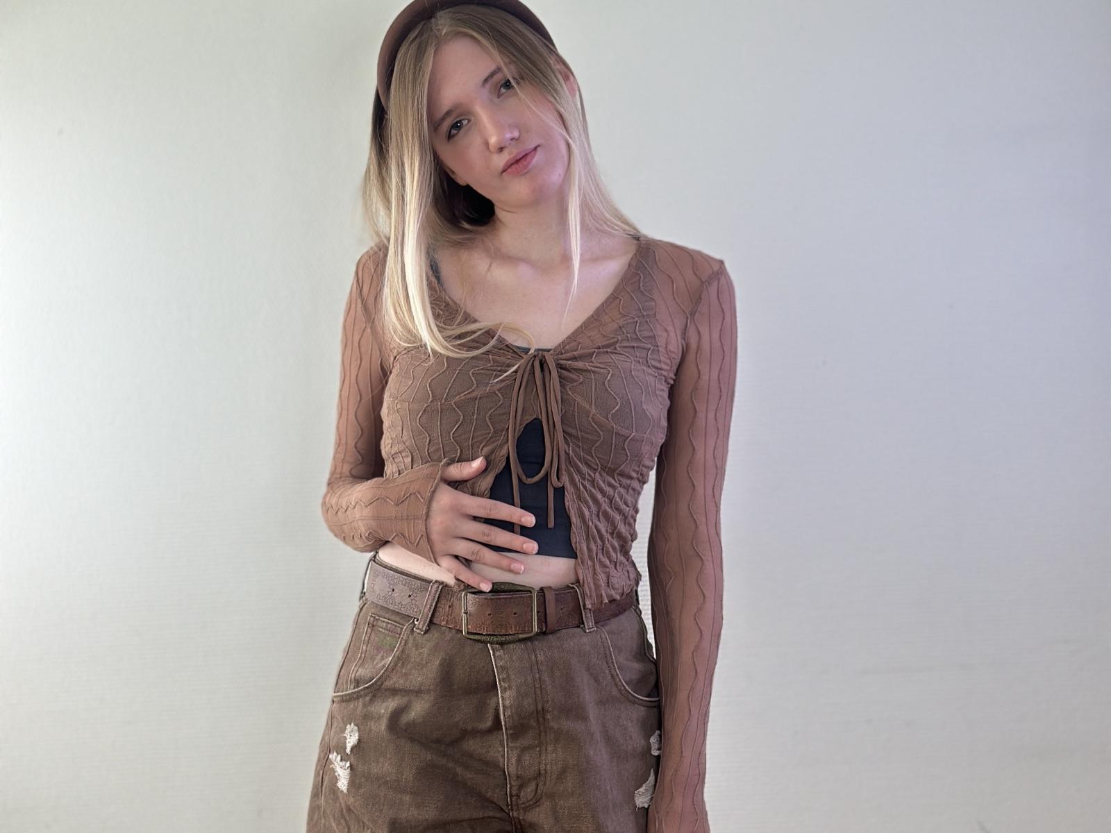 SommerFrymier tiene el cabello rubio, y lleva una blusa marrón transparente de manga larga, pantalones marrones de cintura alta y un sombrero marrón. Sostiene un teléfono negro, con una mano sobre su vientre..