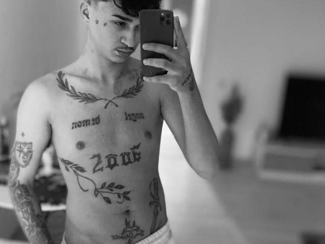IrisIrs with tattoos, shirtless, holding a phone, taking a mirror selfie. IrisIrs has a lean, muscular build, short dark hair, and visible tattoos on chest and arms.