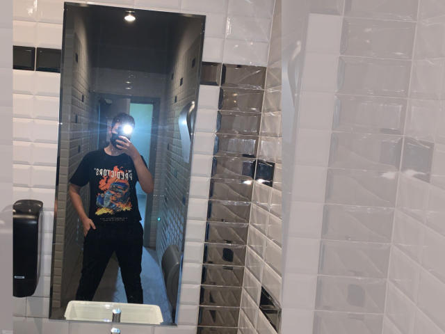 MarcusIg69 stands in a bathroom, wearing a black "Runnin' Down to the Store" t-shirt and black pants, taking a mirror selfie with a camera flash.