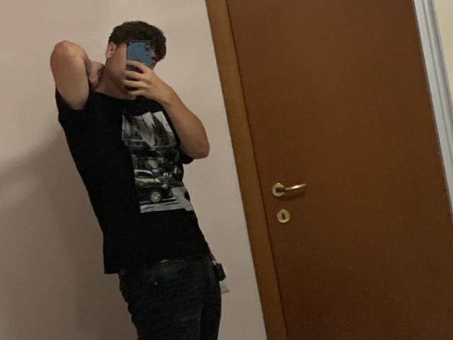 MarcusIg69 steht in einem Spiegel-Selfie, trägt ein schwarzes T-Shirt mit einer Schwarz-Weiß-Grafik, dunkle Jeans und hält ein Handy vor sein Gesicht. Er hebt einen Arm..