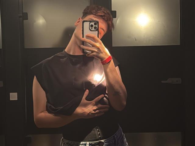 MarcusIg69 with red wristband, black sleeveless shirt, and blue jeans, holding phone in front of face, hand on chest, mirror reflection, visible light reflection, short brown hair.
